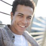 Happy male smiling and looking at camera while leaning on metal wire of fence in sunny day